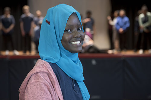 A female performer smiles at the camera while students perform on stage behind her.