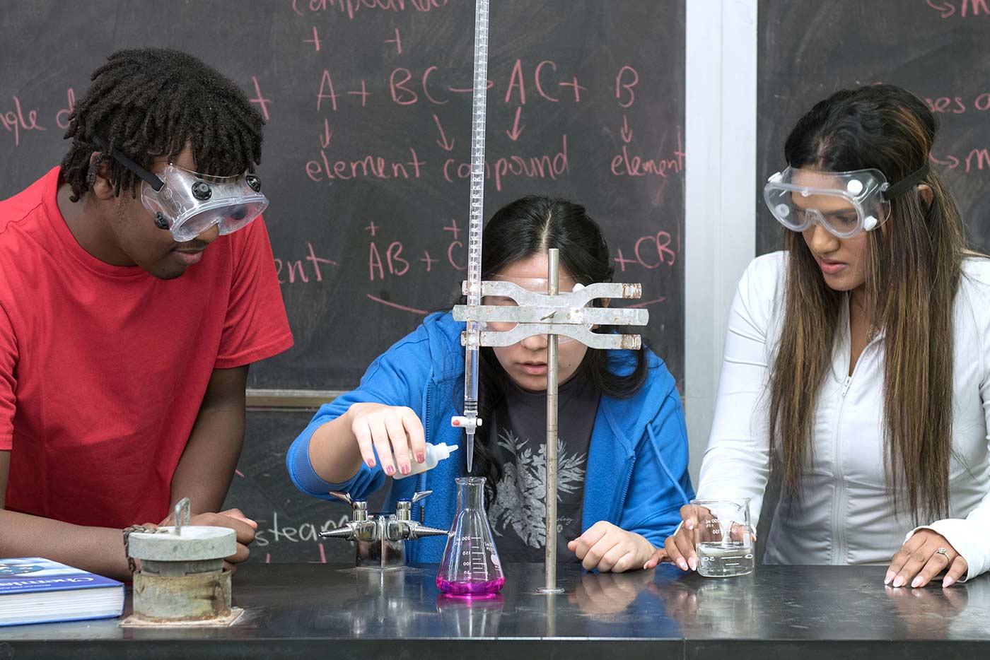 Three students wearing goggles conduct an experiment with beakers and flasks